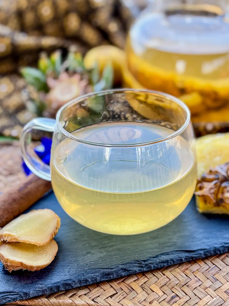 Pineapple tea on a slate tray with fresh ginger slices, a cinnamon stick, pineapple skin, and a teapot in the background.