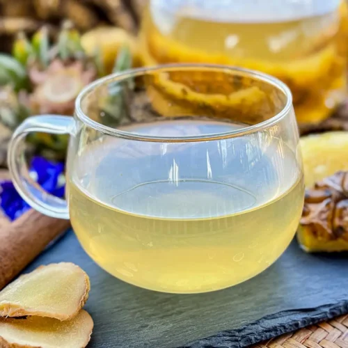 Pineapple tea served with fresh ginger slices, a cinnamon stick, and a teapot filled with pineapple skin and core in the background.