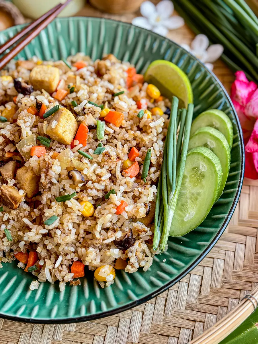A plate of vegetarian Thai fried rice with tofu, carrots, sweet corn, and shiitake mushrooms, garnished with fresh green onions and served with cucumber slices.