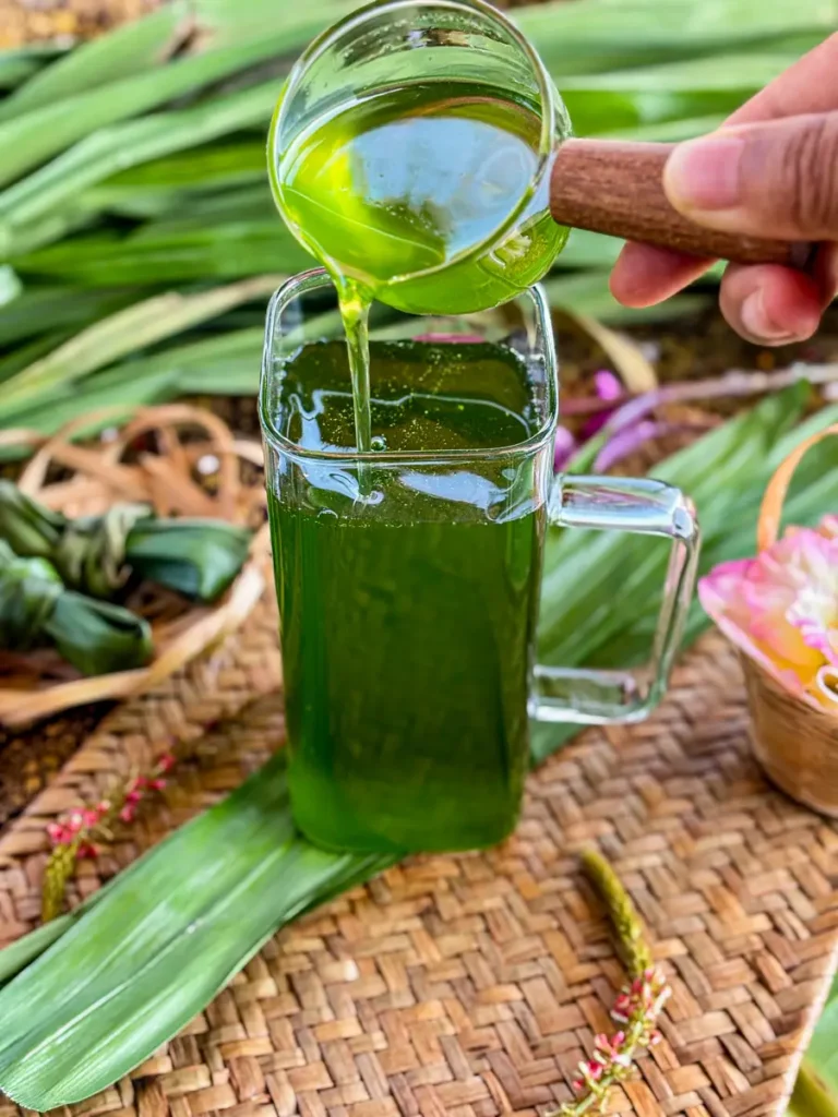 Pouring vibrant green pandan syrup from a small glass cup into a glass jug, surrounded by fresh pandan leaves.