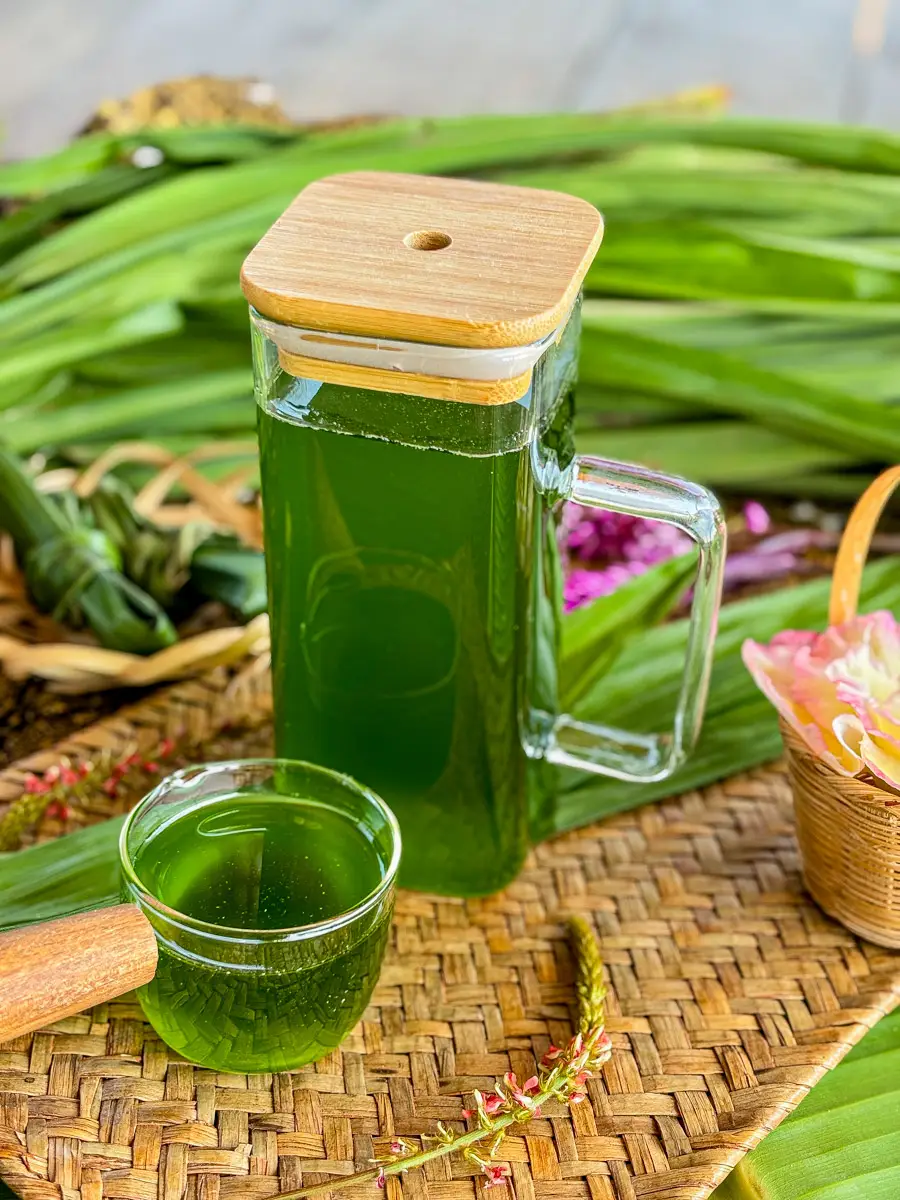 Glass jug of green pandan syrup with a small glass cup, surrounded by pandan leaves and decorative flowers on a woven tray.