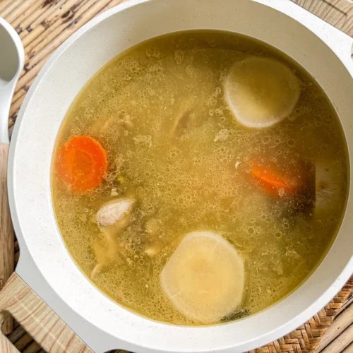 Overhead view of nam stock, Thai broth simmering with carrots, daikon, onion, and bone-in chicken in a white pot