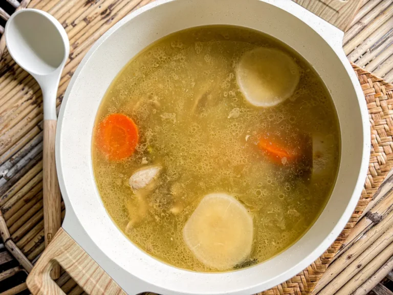 Overhead view of nam stock, Thai broth simmering with carrots, daikon, onion, and bone-in chicken in a white pot