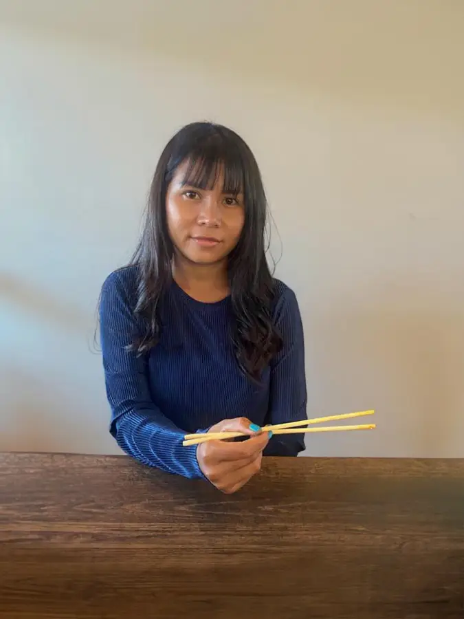 Praew holding chopsticks at a wooden table, wearing a dark blue top.
