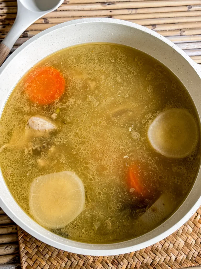 Thai chicken broth with vegetables in a white pot, placed on a woven mat and bamboo surface