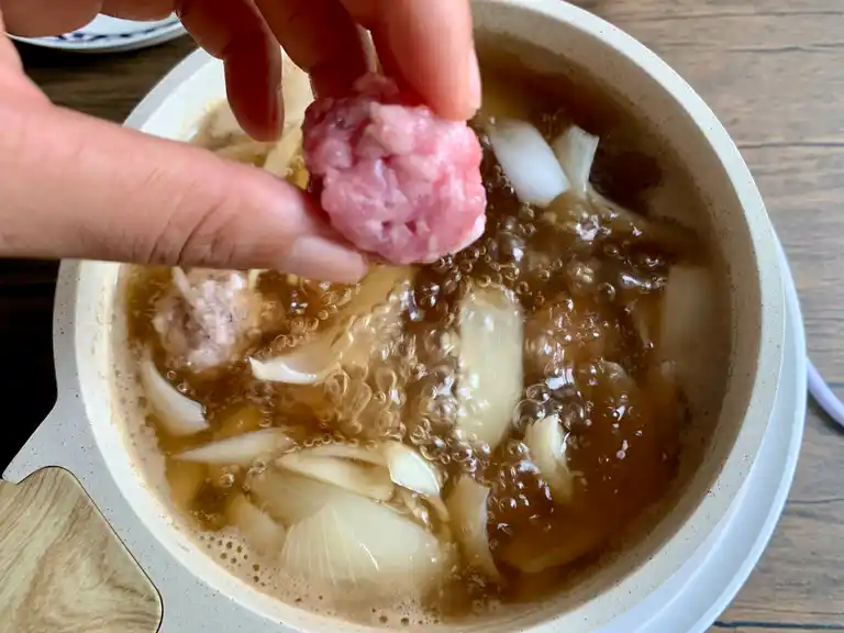 Hand adding raw pork meatball into boiling soup broth.
