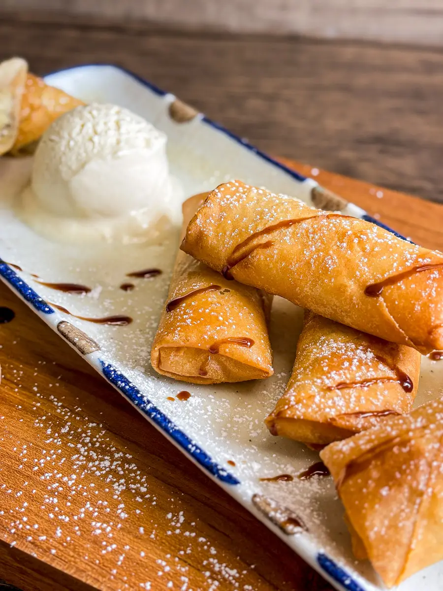 Fried banana spring rolls served with vanilla ice cream, syrup drizzle, and powdered sugar.