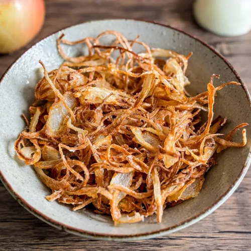 A bowl of crispy fried onions with delicate strands and rich color, served on a rustic wooden surface.