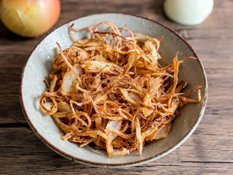 A bowl of crispy fried onions with delicate strands and rich color, served on a rustic wooden surface.