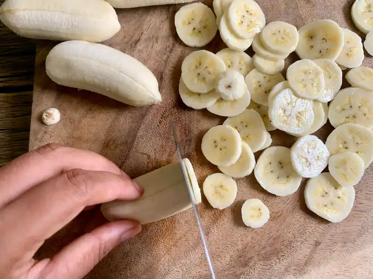 Cutting Thai bananas (kluai nam wa) into thin slices on a wooden cutting board.