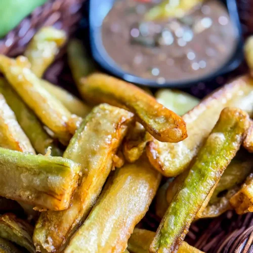 Close-up of golden fried eggplant fries served in a basket with Thai nam prik kapi dipping sauce in the background.
