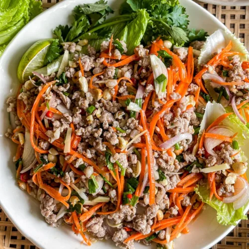 Overhead view of Thai nam sod salad with lime, peanuts, carrots, and fresh herbs, served on a white plate.