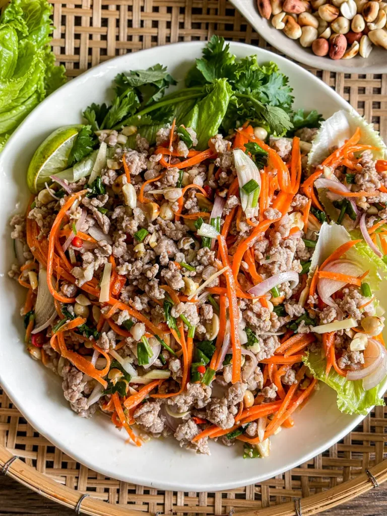 Overhead view of Thai nam sod salad with lime, peanuts, carrots, and fresh herbs, served on a white plate.