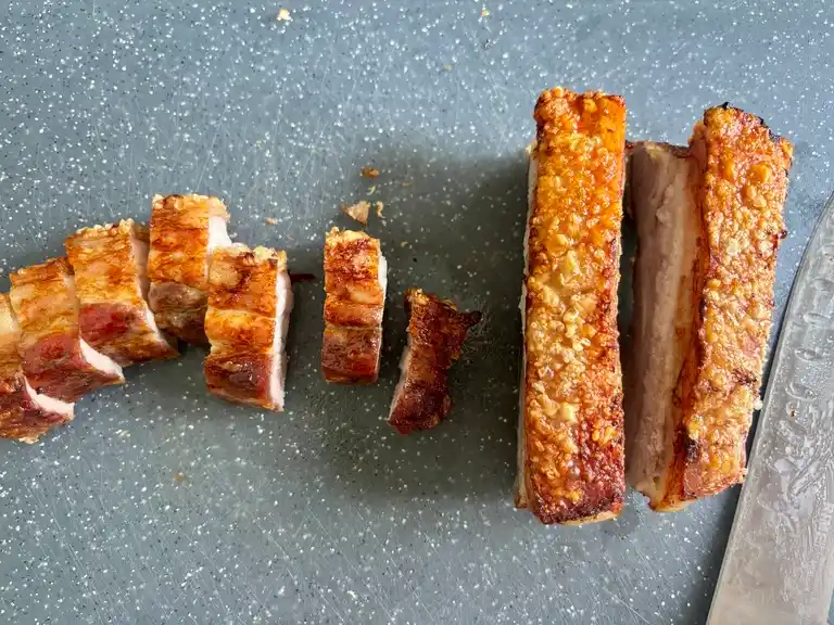 Crispy pork belly being sliced into bite-sized pieces on a cutting board.