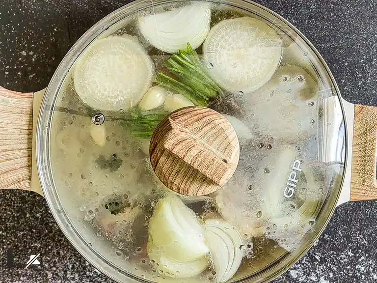 Pot of Thai nam stock simmering with a lid on, filled with daikon, onion, garlic, and herbs..