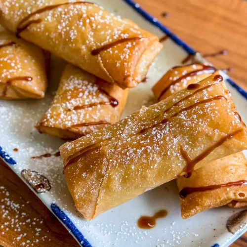 Golden brown Thai fried banana spring rolls drizzled with syrup and dusted with powdered sugar on a rectangular plate.
