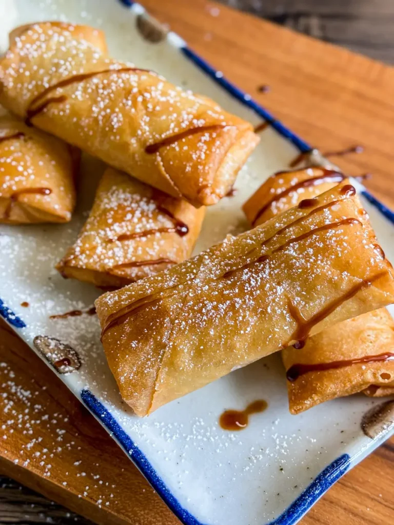 Golden brown Thai fried banana spring rolls drizzled with syrup and dusted with powdered sugar on a rectangular plate.