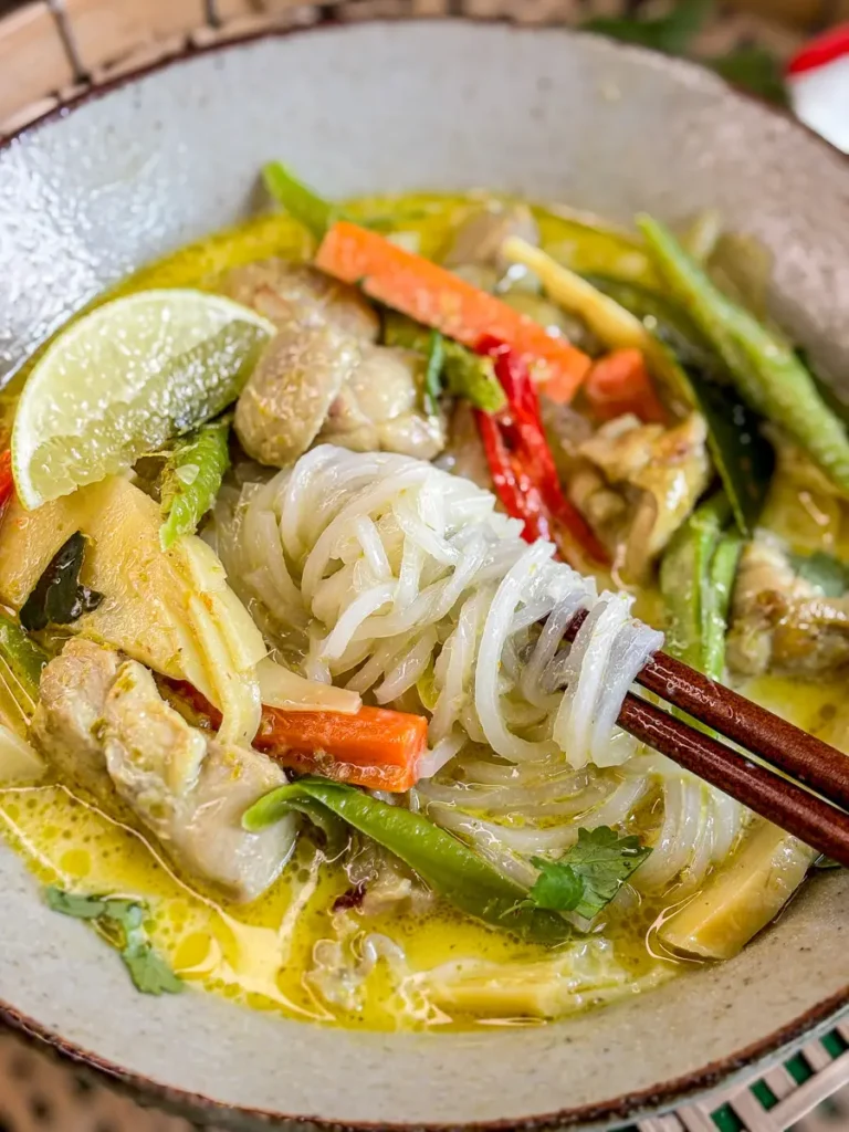 Thai green curry noodle soup with rice noodles, chicken, carrots, and green beans in a creamy coconut milk broth, served with chopsticks.