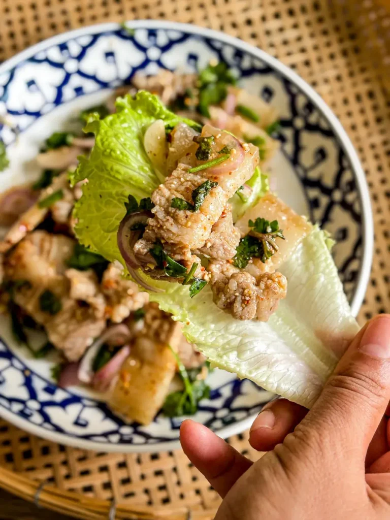 Hand holding a lettuce wrap filled with Thai pork belly larb, fresh herbs, and shallots, ready to eat.