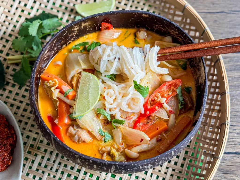 Thai red curry noodle soup with chicken, vegetables, and rice noodles, served in a bowl with lime and fresh herbs.