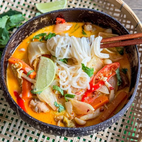 Sopa de macarrão de curry vermelho tailandês com frango, legumes e macarrão de arroz, servido em uma tigela com limão e ervas frescas.