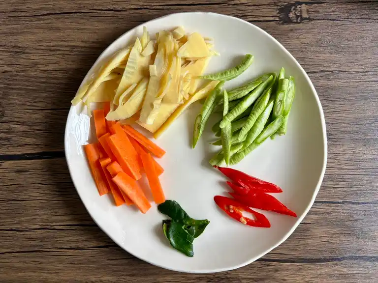 Plate of prepped vegetables for Thai green curry soup including sliced carrots, bamboo shoots, yard long beans, red chili, and kaffir lime leaves.