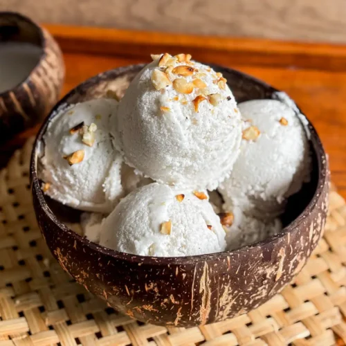 Thai coconut milk ice cream with chopped peanuts in a coconut shell bowl, styled on a woven bamboo mat with coconut milk on the side.