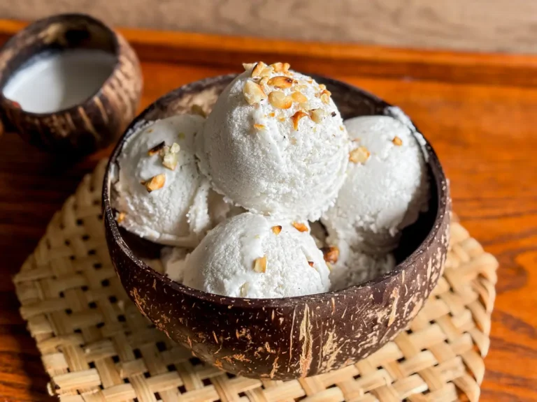 Thai coconut milk ice cream with chopped peanuts in a coconut shell bowl, styled on a woven bamboo mat with coconut milk on the side.