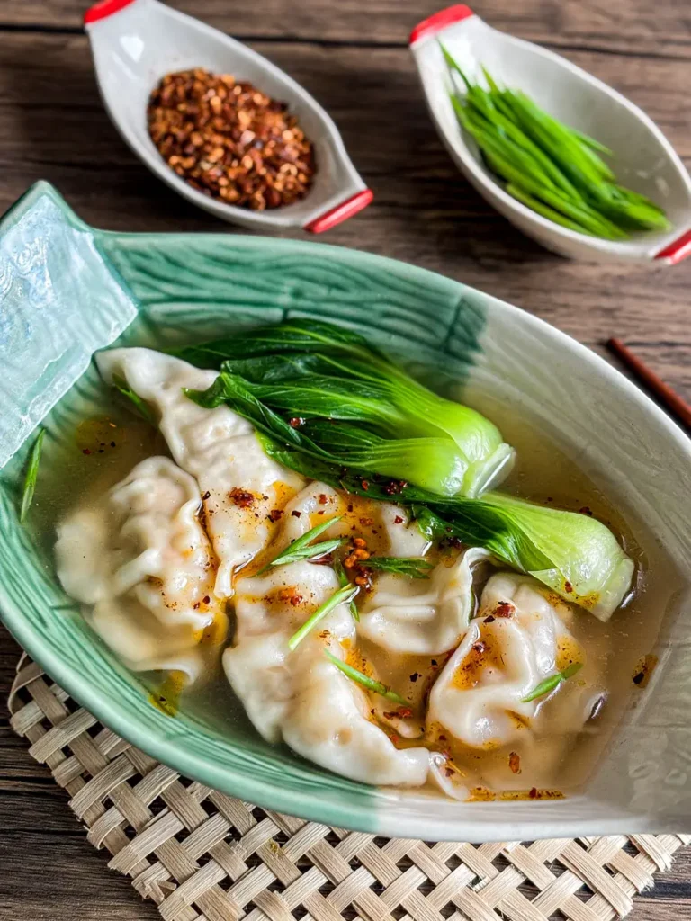 Homemade Thai wonton soup served in a green bowl with bok choy and chili flakes on top.
