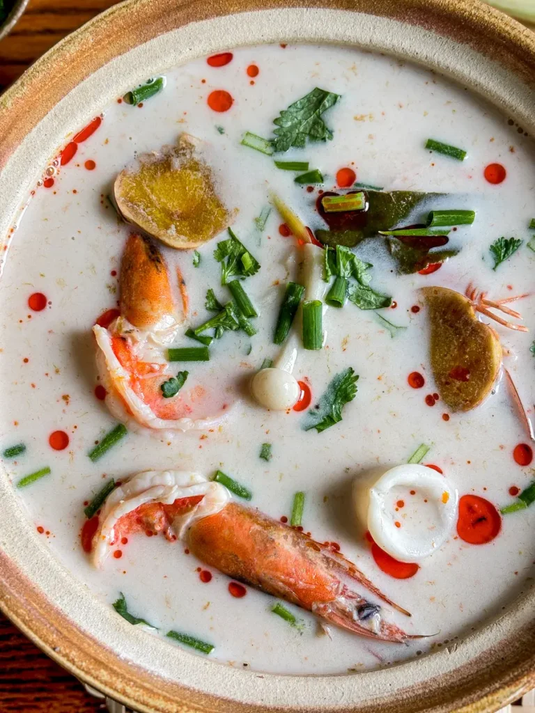Close-up of Thai seafood tom kha soup with shrimp heads, squid rings, lemongrass, and fresh herbs in a coconut milk broth.