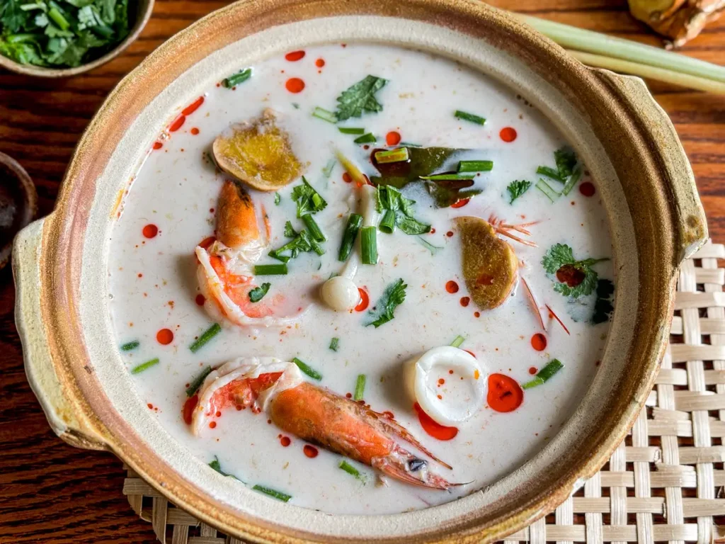 Tom kha talay in a clay bowl, showing shrimp, squid, galangal slices, kaffir lime leaves, and chili oil on creamy coconut broth.