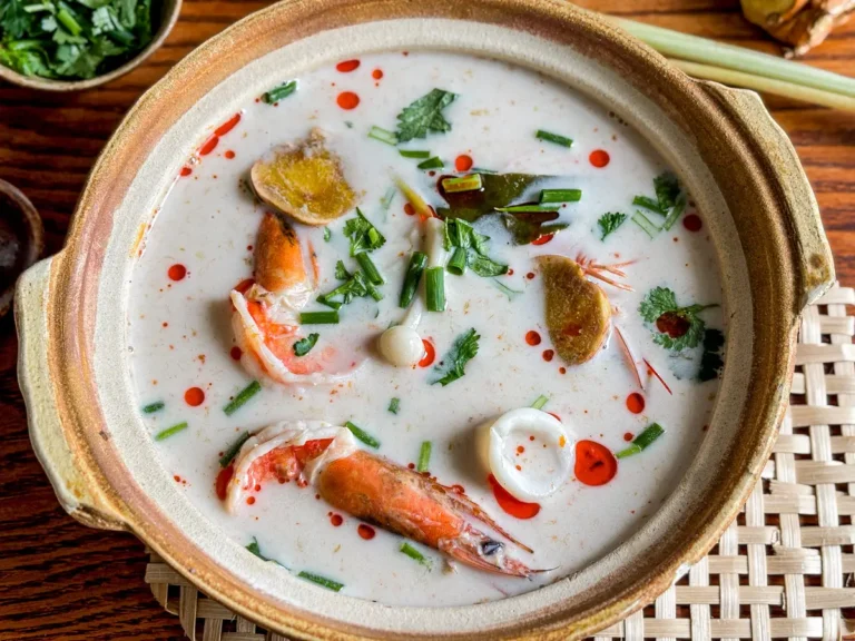 Tom kha talay in a clay bowl, showing shrimp, squid, galangal slices, kaffir lime leaves, and chili oil on creamy coconut broth.