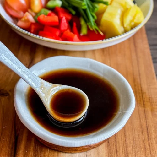 Sweet and sour stir-fry sauce in a small white bowl with a ceramic spoon, served on a wooden board next to fresh vegetables.