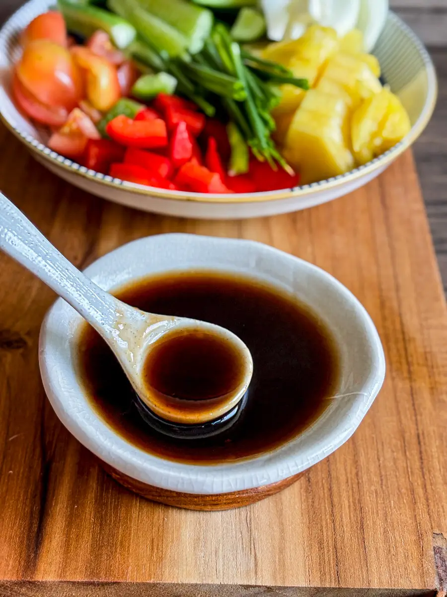 Sweet and sour stir-fry sauce in a small white bowl with a ceramic spoon, served on a wooden board next to fresh vegetables.