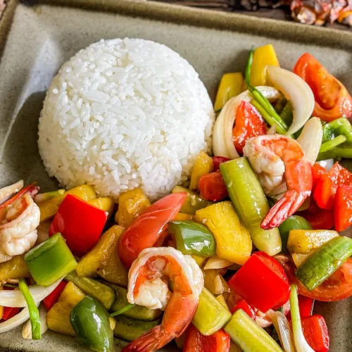 Thai sweet and sour shrimp stir-fry with colorful vegetables and pineapple, served with a dome of jasmine rice on a ceramic plate.
