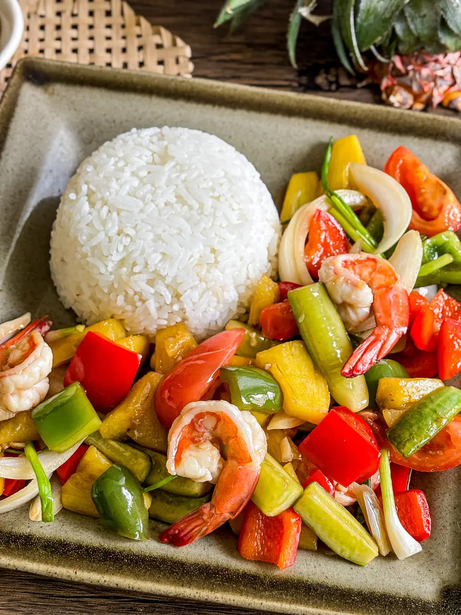 Thai sweet and sour shrimp stir-fry with colorful vegetables and pineapple, served with a dome of jasmine rice on a ceramic plate.