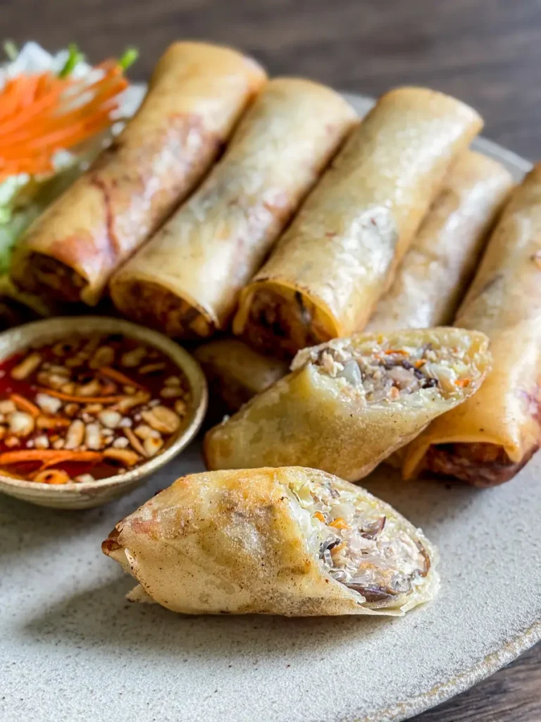 Close-up of crispy fried spring rolls on a plate with one cut open to show the savory pork and vegetable filling, served with a Thai dipping sauce on the side.