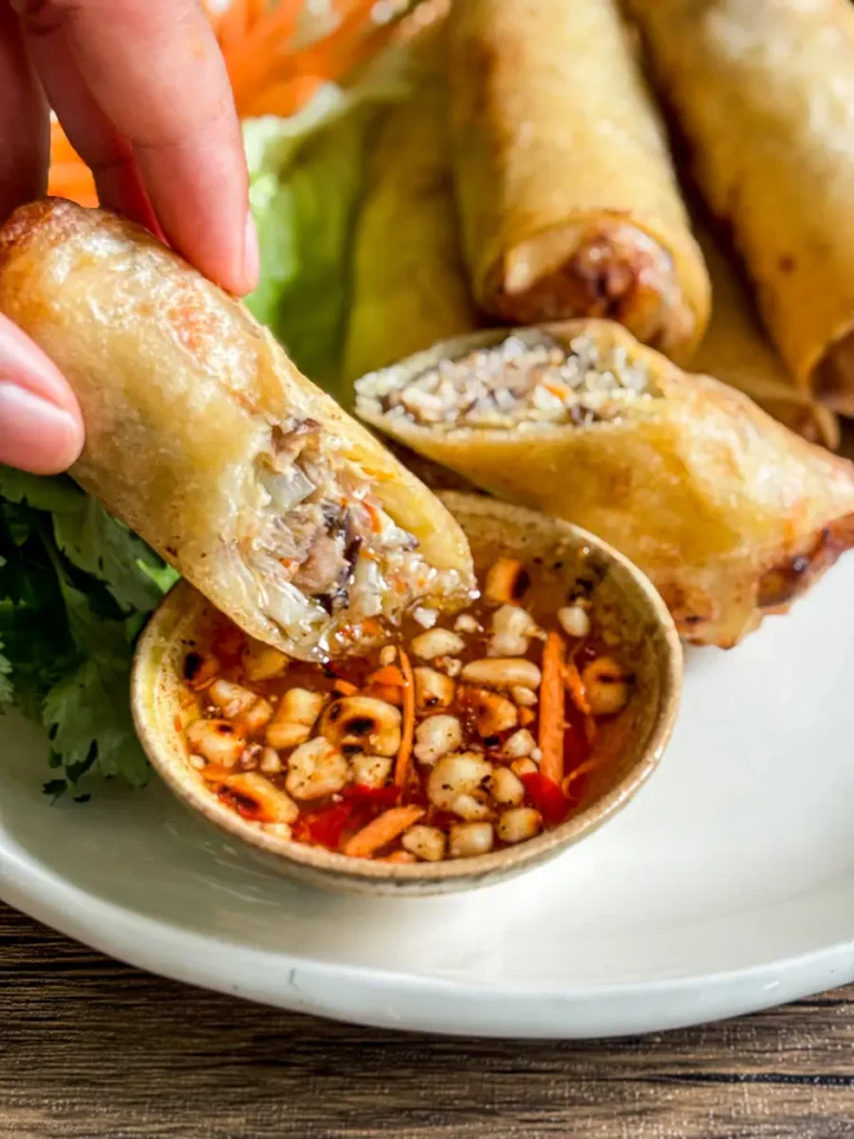 A hand dipping a crispy spring roll into Thai dipping sauce for spring rolls with chopped peanuts, shredded carrot, and sliced chili.
