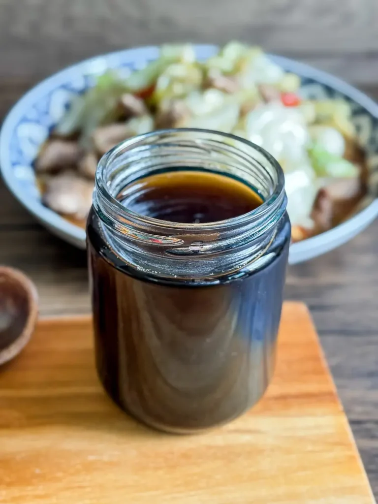 Homemade Thai stir fry sauce in a glass jar with a plate of stir-fried vegetables and beef in the background.