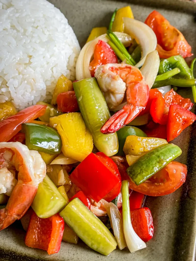 Close-up of Thai sweet and sour shrimp with pineapple, bell peppers, cucumber, onion, and tomato served with steamed jasmine rice.