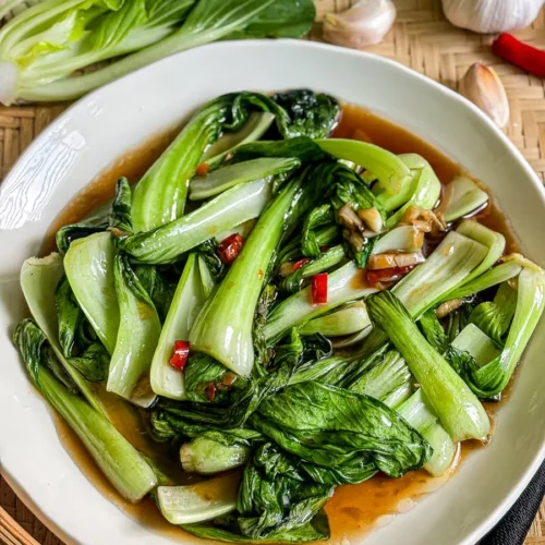 Garlic bok choy stir-fry served in a light brown sauce, with fresh bok choy, garlic cloves, and Thai chili in the background.