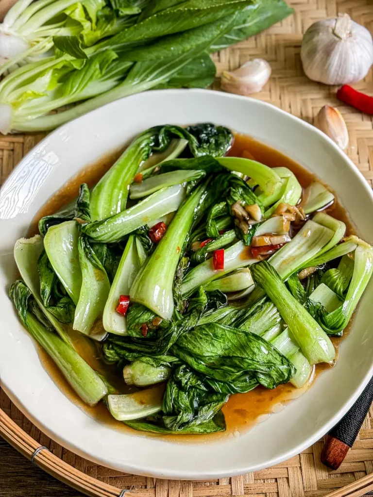 Garlic bok choy stir-fry served in a light brown sauce, with fresh bok choy, garlic cloves, and Thai chili in the background.