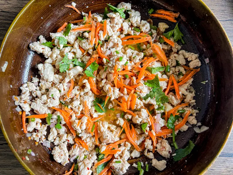 Cooked minced chicken, shredded carrots, and chopped herbs tossed in dressing in a large mixing bowl.