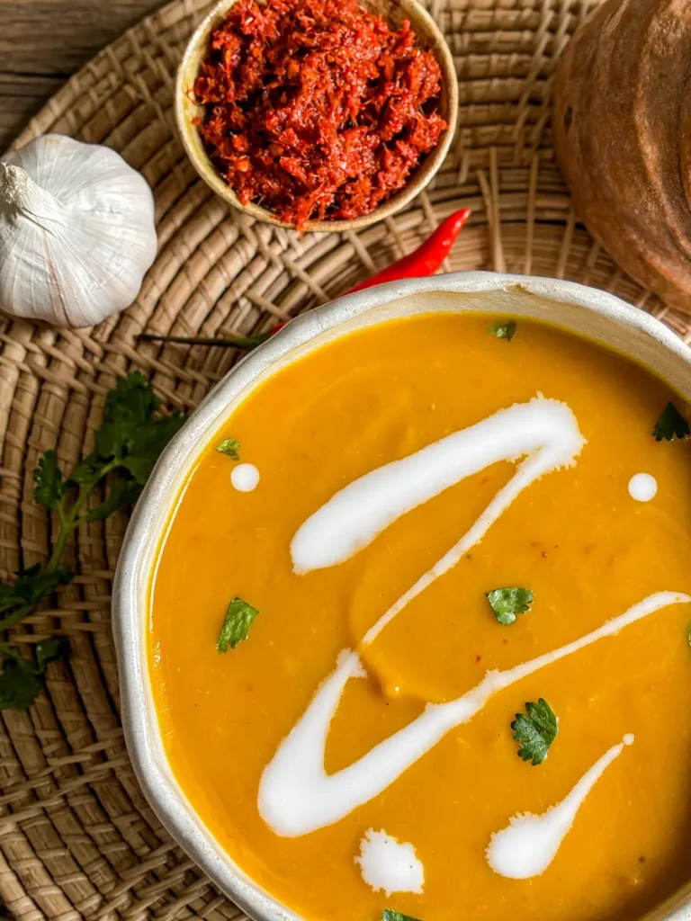 Sopa cremosa tailandesa de coco e abóbora com coco ralado e coentro, rodeada de pasta de curry vermelho, alho, pimenta e ervas em uma esteira tecida.