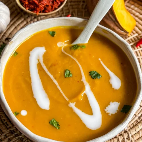 Sopa de abóbora tailandesa coberta com creme de coco e coentro, servida em uma tigela de cerâmica com pasta de curry vermelho tailandês, alho fresco e abóbora como acompanhamento.