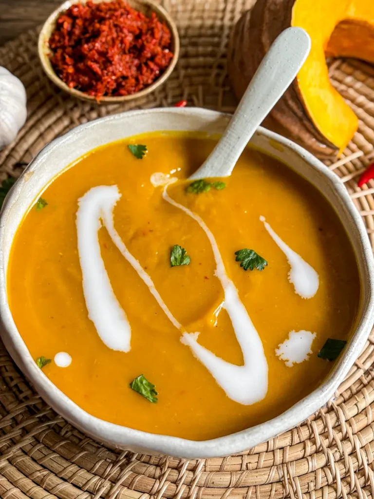 Thai pumpkin soup topped with coconut cream and cilantro, served in a ceramic bowl with Thai red curry paste, fresh garlic, and pumpkin on the side.