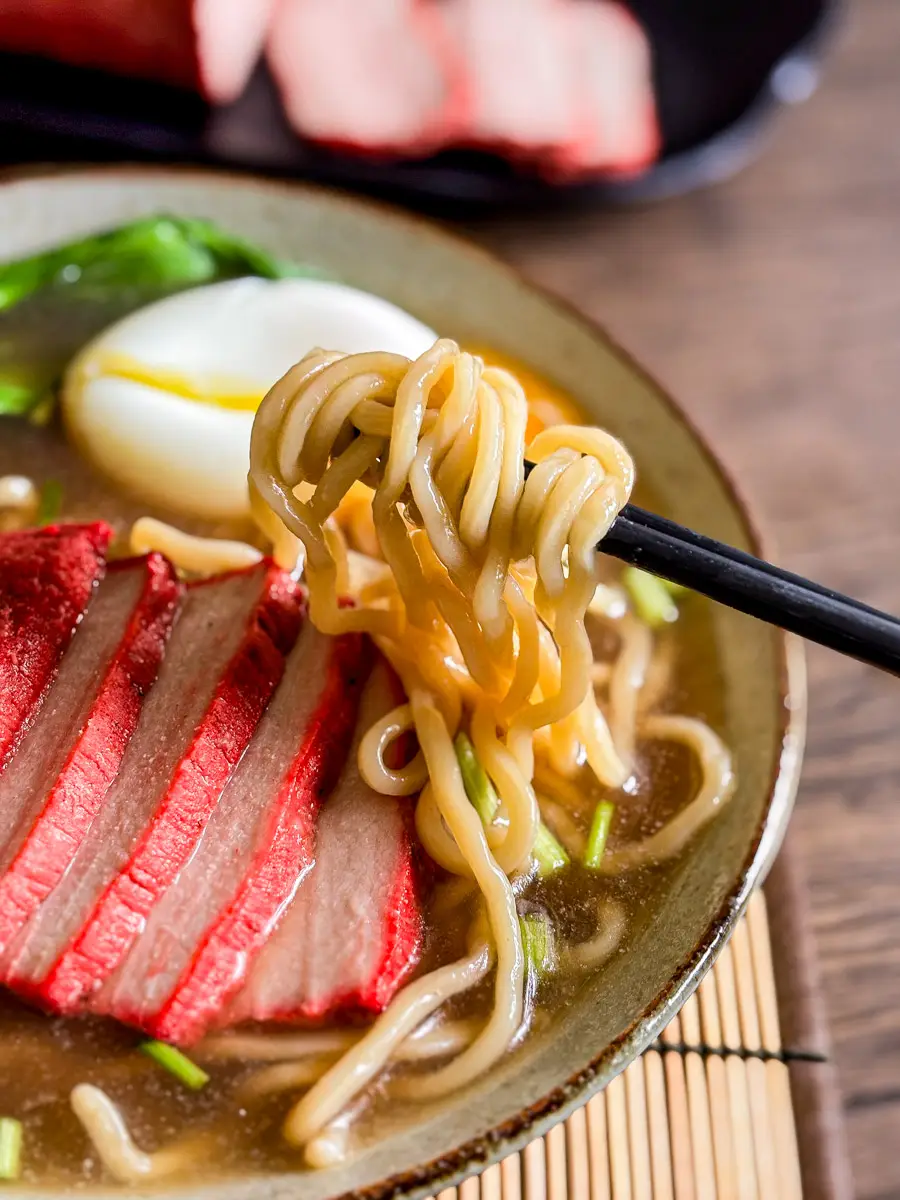 Chopsticks lifting ramen noodles from a bowl of char siu ramen with BBQ pork, egg, and greens.