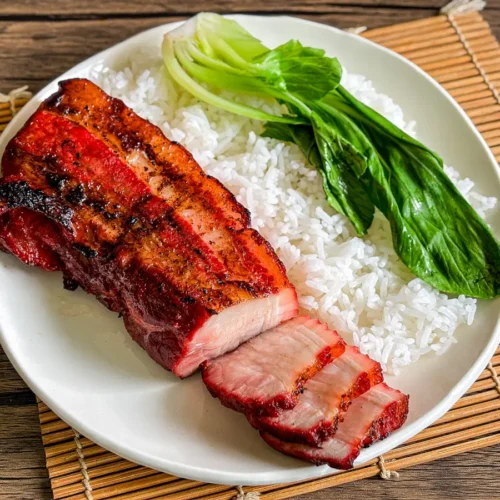 Overhead view of char siu pork belly with rice and bok choy on a white plate and bamboo placemat.
