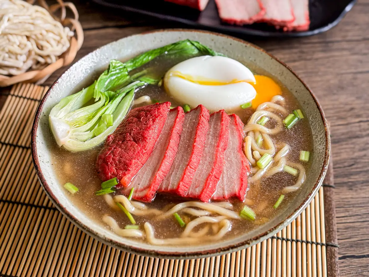 Pork char siu ramen served with bok choy, ramen noodles, soft egg, and red BBQ pork, with extra sliced pork in the background.