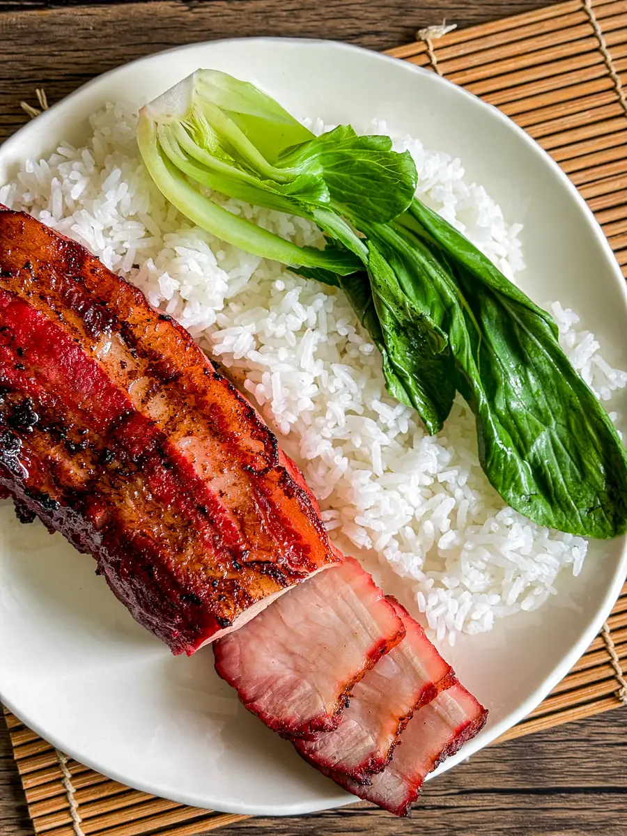 Close-up of juicy char siu pork belly slices on rice with fresh bok choy, served on a white plate.